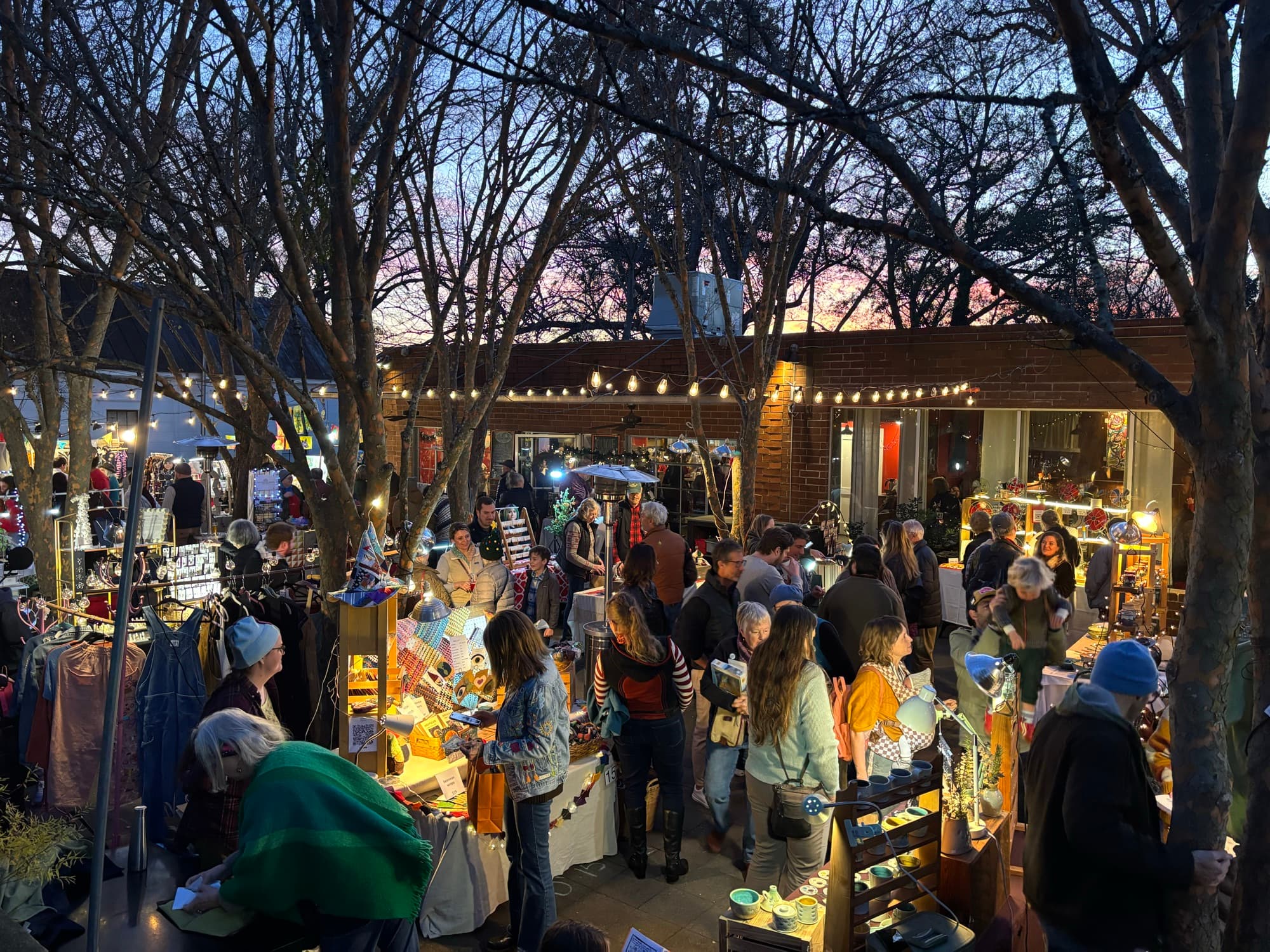 An overhead view of the tree-lit courtyard packed with shoppers browsing artist booths at dusk