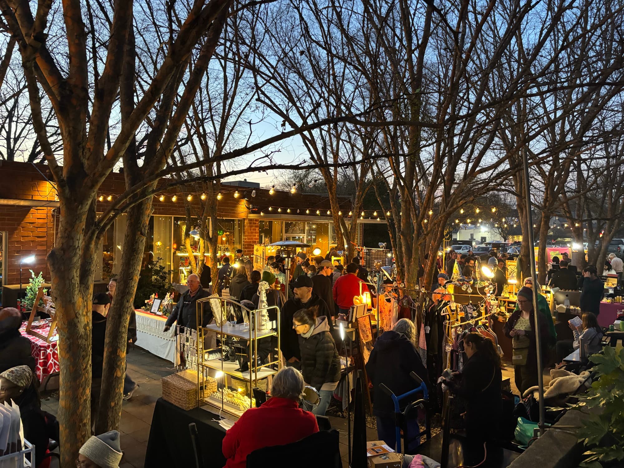Shoppers browsing glowing artist booths among string-lit trees in the courtyard at blue hour